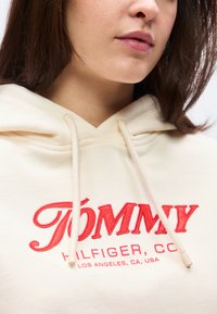 Κοντινό πλάνο ενός ατόμου που φοράει εκρού φούτερ με κουκούλα, με κόκκινα κεντημένα γράμματα «Tommy Hilfiger Co.» και «Los Angeles, CA, USA» στο στήθος.