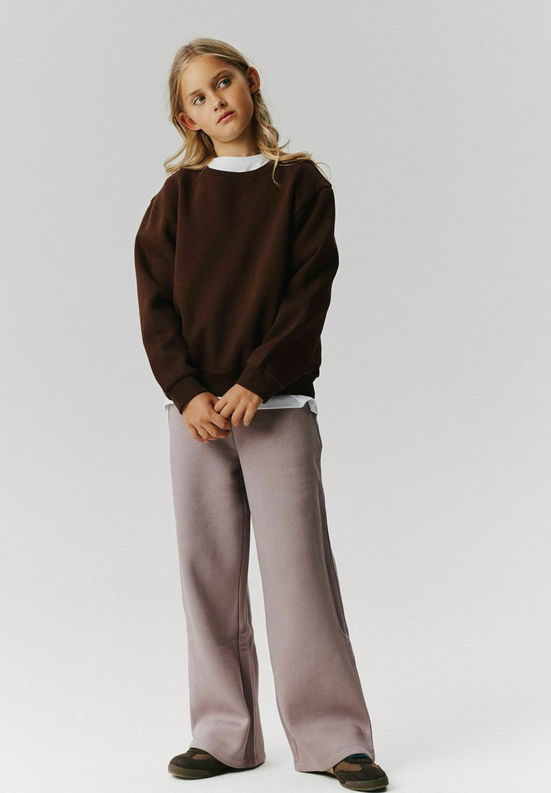 Jeune personne aux longs cheveux portant un sweat-shirt marron foncé, un pantalon large gris clair et des chaussures foncées, debout devant un fond uni.