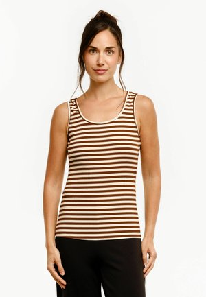 Femme aux cheveux foncés attachés, portant un haut sans manches rayé marron et beige et un pantalon noir, debout devant un fond blanc.