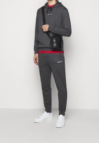 Felpa grigia con tasca frontale, abbinata a pantaloni jogger coordinati. Sottot-shirt rossa visibile, accompagnata da una piccola borsa a tracolla nera e sneakers bianche.