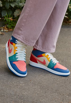 Osoba nosząca kolorowe trampki Nike z koralowymi, niebieskimi, turkusowymi, czerwonymi, żółtymi i białymi akcentami na szarym chodniku na zewnątrz.