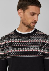 Mężczyzna w czarnym swetrze z okrągłym dekoltem, z czerwonym, białym i szarym wzorem fair isle, patrzący w dół i na bok na tle jednolitym.