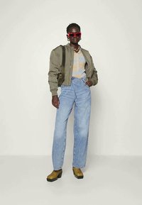 Oliivroheline pommijakk, beež graafiline T-särk, helesinised teksad, tursked sinepikollased puhtad ja punased prillid, seistes neutraalse tausta ees.