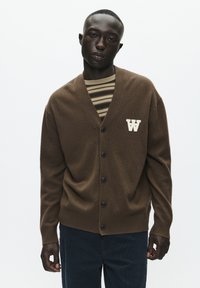 Brązowy kardigan z dekoltem w kształcie litery V, zapinany na guziki, z prążkowanym wykończeniem. Ma kremowe logo „W” i jest noszony na paskowanym T-shircie.