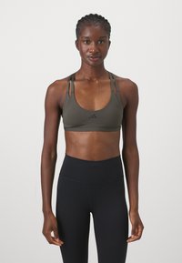 Top sportivo di colore verde oliva scuro con scollatura rotonda, sottili spalline regolabili e un piccolo logo nero sul davanti. Abbinato a leggings neri a vita alta.