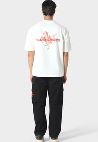Camiseta blanca con estampado gráfico en rojo en la parte posterior, que presenta el texto "PAS DE MONACO" y una imagen estilizada. Pantalones cargo negros con bolsillos laterales.