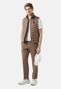 Hnědá prošívaná vesta s vertikálními švy, bílá polokošile, hnědé kalhoty a bílá čepice. Model má na sobě tenisky v neutrálních tónech.