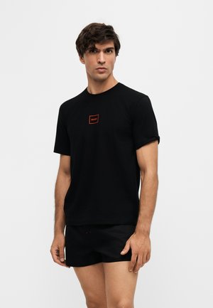 Junger Mann mit dunklen Haaren, der ein schwarzes T-Shirt mit kleinem rotem quadratischem Logo und schwarze Shorts trägt, steht vor einem schlichten hellen Hintergrund.