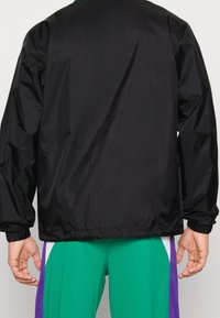 Zwarte windbreaker jas met elastische boorden en zoom, gladde textuur, lichtgewicht materiaal. Achteraanzicht; gecombineerd met groene en paarse broek.