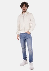 Giacca leggera color crema con zip e colletto alto, indossata sopra una camicia bianca e jeans azzurri chiari, abbinata a sneakers bianche.