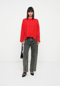 Rode cropped trui met verlaagde schouders, gecombineerd met hooggerande, loszittende olijfgroene jeans en zwarte pumps met spitse neuzen. Draagt een zwarte handtas.