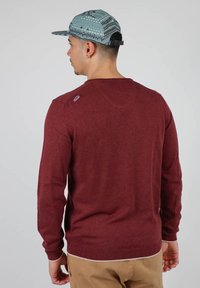 Pull en tricot bordeaux à manches longues, avec des poignets et un ourlet côtelés, arborant un petit logo sur l'épaule. Porté avec un pantalon beige et une casquette à motifs.