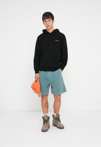 Black hoodie with front pocket and logo. Light blue denim shorts. Gray boots with laces. Orange cap held in hand. Clean, minimal background.