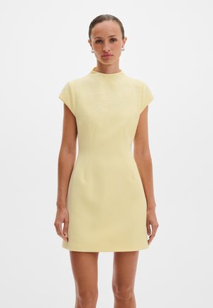 Kvinne iført en lys gul, kortermet kjole til knærne med subtil blomsterbroderi på øvre del av brystet, foran en hvit bakgrunn.