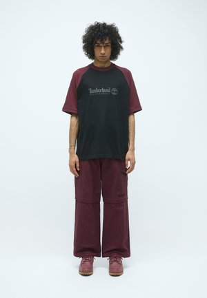 Black and burgundy short-sleeve shirt with logo, paired with burgundy wide-leg pants and matching burgundy shoes with textured soles.