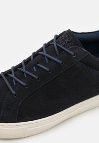 Adidași din suede albastru marin cu o suprafață texturată, având șireturi asortate și o talpă din cauciuc alb cu detalii de cusătură discrete.