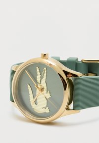 Kulaté hodinky se zlatým rámečkem, zeleným silikonovým páskem a zeleným ciferníkem s vyvýšeným zlatým logem krokodýla a zlatými ručičkami.