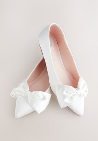 Zapatos planos blancos de punta afilada con grandes lazos de satén en la parte delantera y forro interior rosa claro, colocados sobre una superficie lisa y clara.