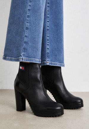 Bottines noires à talons hauts avec semelles robustes portées avec un jean bleu à jambes larges sur un fond neutre.