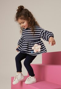 Teksaskandiga meresinine ja valge ülaosa, millel on laienev äär ja luksuslik lilleelement, koos meresiniste leggingsite ja valgete kõrgemate spordijalatsitega.