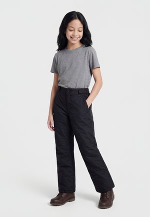 Ung person med langt svart hår, iført en grå kortermet skjorte, svarte bukser og brune støvler, stående med en hånd i lommen.