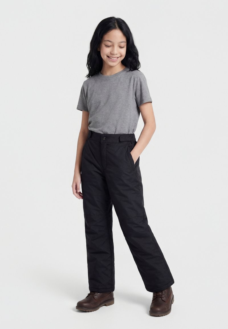 Mladý člověk s dlouhými černými vlasy, oblečený ve šedém tričku s krátkým rukávem, černých kalhotách a hnědých botách, stojící s jednou rukou v kapse.