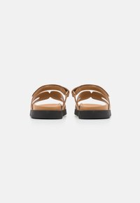 Braune Leder-Sandalen mit Ausschnittdesign, flacher schwarzer Sohle und glattem hellbraunem Fußbett. Offene Rückseite mit dekorativen Riemen für einen sicheren Sitz.