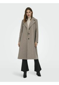 Femme portant un long manteau beige avec des boutons noirs, un pull col roulé crème, un pantalon noir et des bottes à talons noires, se tenant face à l'avant.