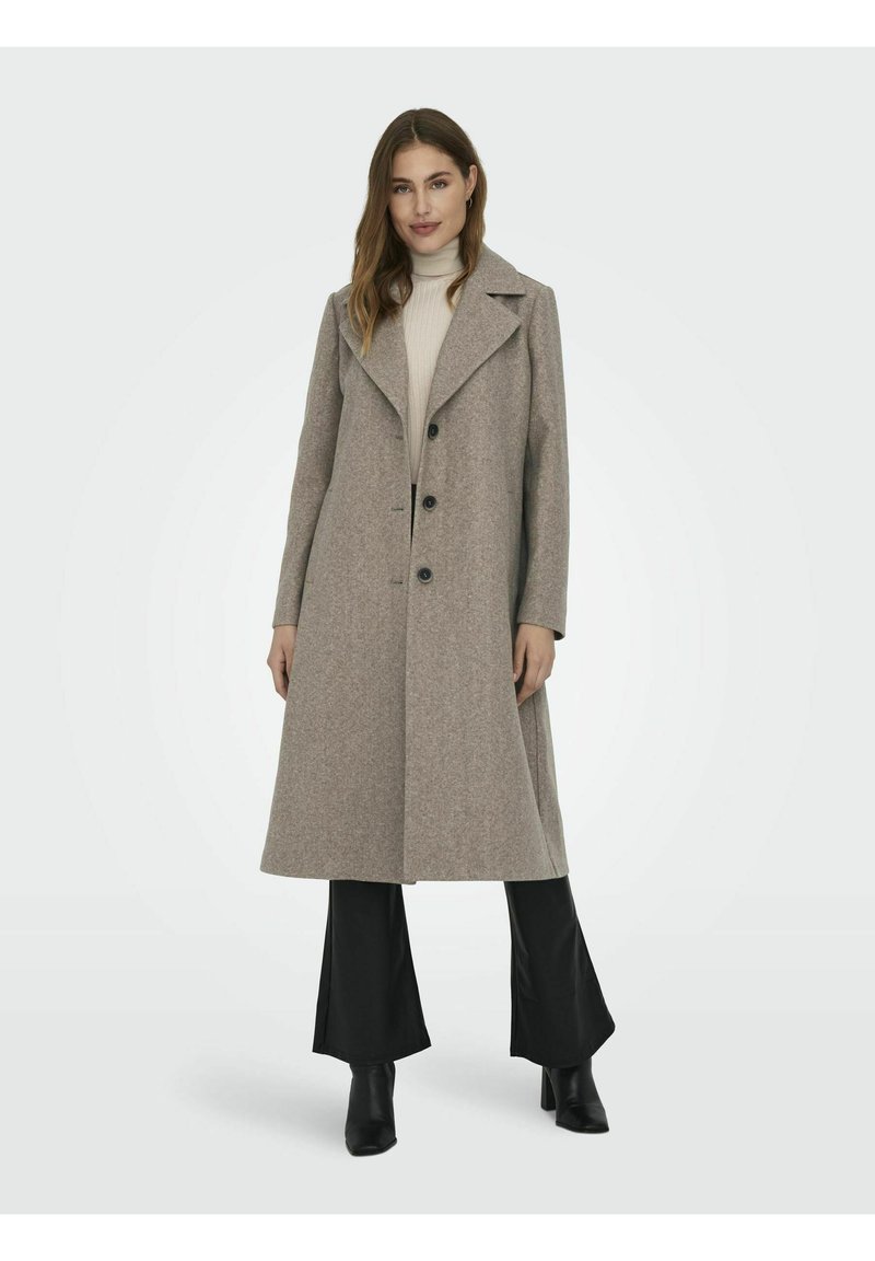 Femme portant un long manteau beige avec des boutons noirs, un pull col roulé crème, un pantalon noir et des bottes à talons noires, se tenant face à l'avant.