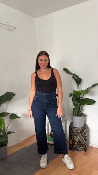 Bodysuit preto sem mangas, calças de ganga escuras de cintura alta com detalhes em dourado, ténis brancos, em pé sobre um piso de madeira perto de plantas de interior.