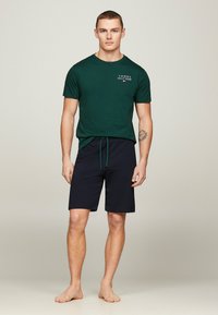 T-shirt verde in cotone con maniche corte e piccolo logo; pantaloni corti blu navy con vita regolabile, vestibilità ampia e orlo dritto, indossati a piedi nudi.