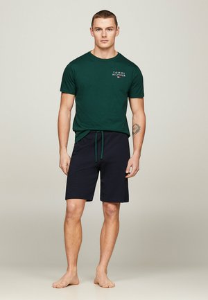 T-shirt verde in cotone con maniche corte e piccolo logo; pantaloni corti blu navy con vita regolabile, vestibilità ampia e orlo dritto, indossati a piedi nudi.