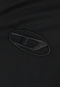 Crni materijal s izvezenim logotipom u sličnoj nijansi. Dizajn je jednostavan, s glatkom teksturom i bez dodatnih uzoraka ili naglasaka.