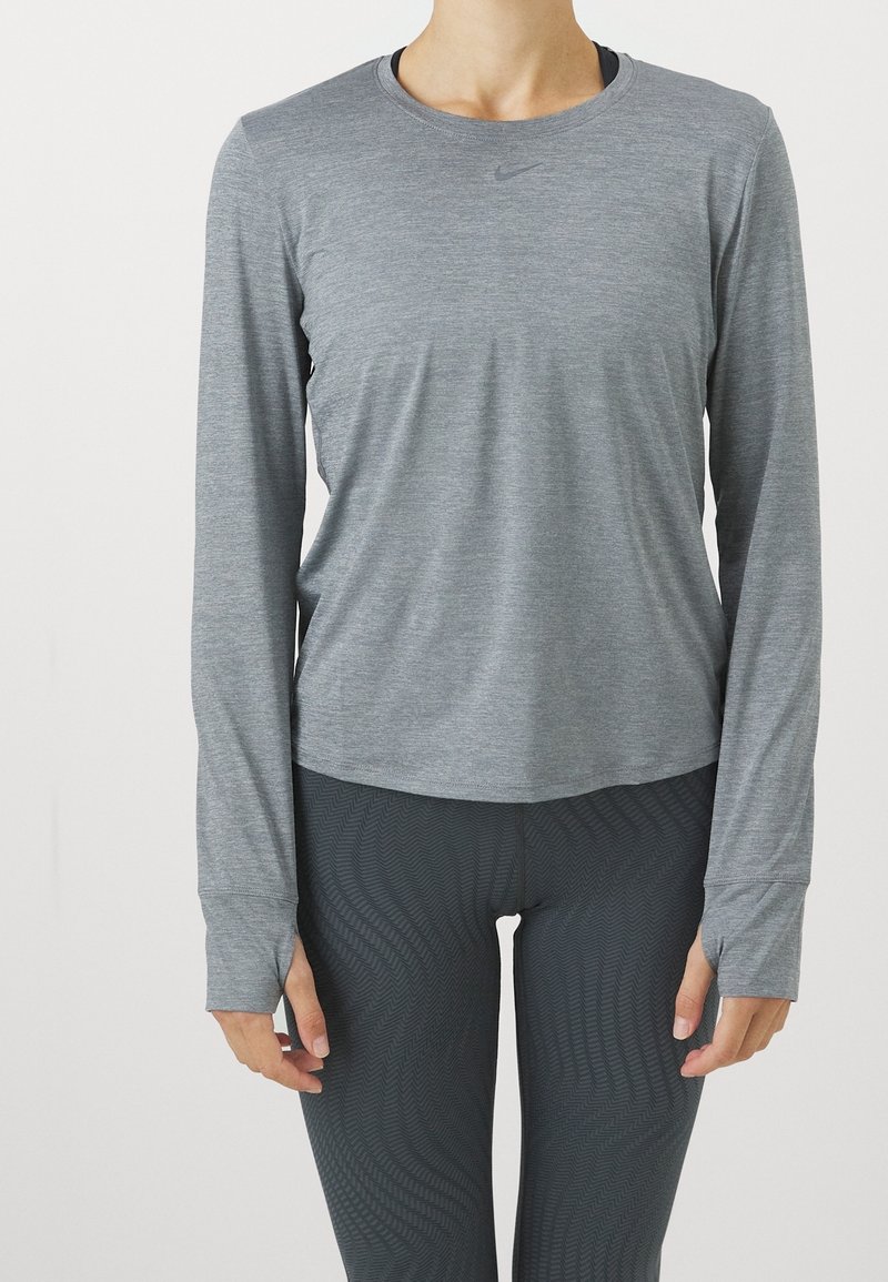 Harmaa pitkähihaurheilupaita, jossa on pyöreä pääntie ja peukalonreiät, yhdistetty tummiin kuvioituihin legginsseihin, valmistettu joustavasta, kosteutta siirtävästä materiaalista.