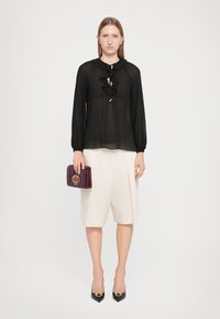Transparante zwarte blouse met een ruches halslijn, gecombineerd met crèmekleurige shorts. Het model houdt een bordeauxrood handtas met reliëflogo vast en draagt zwarte puntige hakken.