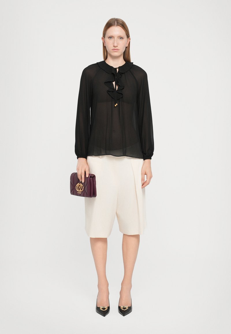 Transparante zwarte blouse met een ruches halslijn, gecombineerd met crèmekleurige shorts. Het model houdt een bordeauxrood handtas met reliëflogo vast en draagt zwarte puntige hakken.