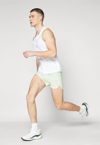 Vit sporttopp, ljusgröna shorts med meshstruktur och vita sneakers med gröna accenter. Ämnet springer.
