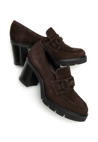 Mocassins à talons en daim marron avec un bout carré, présentant une sangle décorative et une semelle en caoutchouc noir épaisse et texturée.