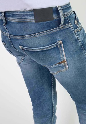 Jeans in denim azzurro chiaro con vestibilità slim. Caratteristiche includono una toppa in pelle nera, due tasche posteriori e dettagli di cucitura distintivi.
