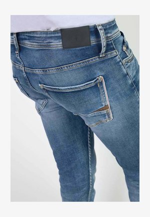 Jeans in denim azzurro chiaro con vestibilità slim. Caratteristiche includono una toppa in pelle nera, due tasche posteriori e dettagli di cucitura distintivi.