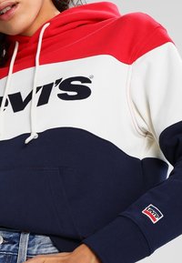 Sweat à capuche bicolore rouge, blanc et marine en mélange de coton, avec poche avant, capuche ajustable par cordon et logo brodé sur la poitrine.