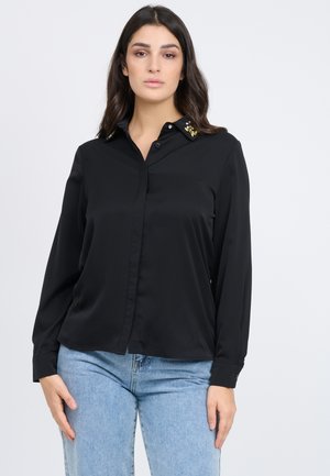 Camisa negra de botones con mangas largas, con cuello tipo solapa y detalles decorativos en el cuello. Tela suave con un corte relajado.