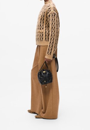Femme portant un pull en maille torsadée beige avec des boutons dorés, un pantalon pattes d'éléphant marron, des chaussures à motif léopard, et tenant un sac à main matelassé noir.