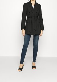 Blazer negro de doble botonadura con cinturón, con bolsillos laterales, usado sobre una blusa blanca y combinado con jeans ajustados azul oscuro y sandalias de tacón negras.