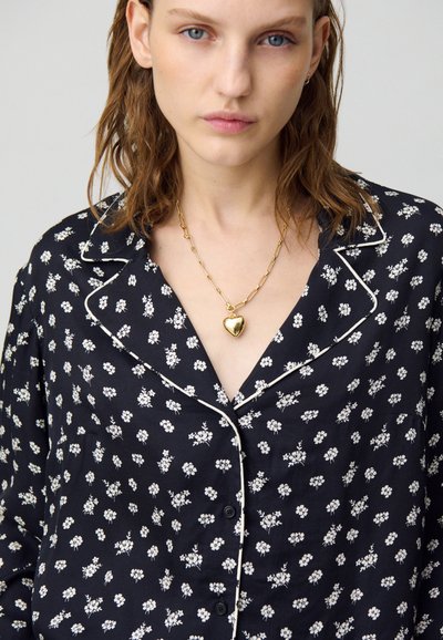 Camicia da pigiama nera con motivo floreale e accenti bianchi. Presenta un colletto a scollo a V, chiusura con bottoni e tessuto leggero. Collana a forma di cuore in oro.