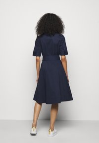 En kvinna med lockigt hår har på sig en marinblå klänning med knyt i midjan, i kombination med vita sneakers med gulddetaljer, vänd bort från kameran.