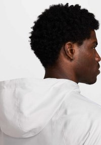 Witte hoodiejack met een gladde textuur, zichtbare stiksels en een trekkoord. Donker, krullend haar en een oorbel zichtbaar in het zijprofiel.