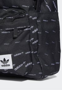 Svart Adidas-ryggsäck med upprepade vita Adidas-logotyper, framficka med dragkedja, och en sydd vit varumärkesetikett på texturerad tyg.