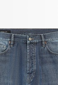 Modré džínové kalhoty s předními kapsami, kovovým zapínáním na knoflík, poutky na pásek a viditelnou štítkem značky Massimo Dutti na vnitřním pasu.