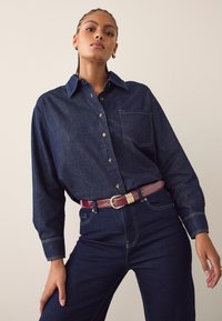 Jeanshemd in Dunkelblau mit durchgehendem Knopfleistenverschluss, langen Ärmeln und einer Brusttasche. Kombiniert mit hochtaillierter dunkler Jeanshose und einem burgunderroten Gürtel.